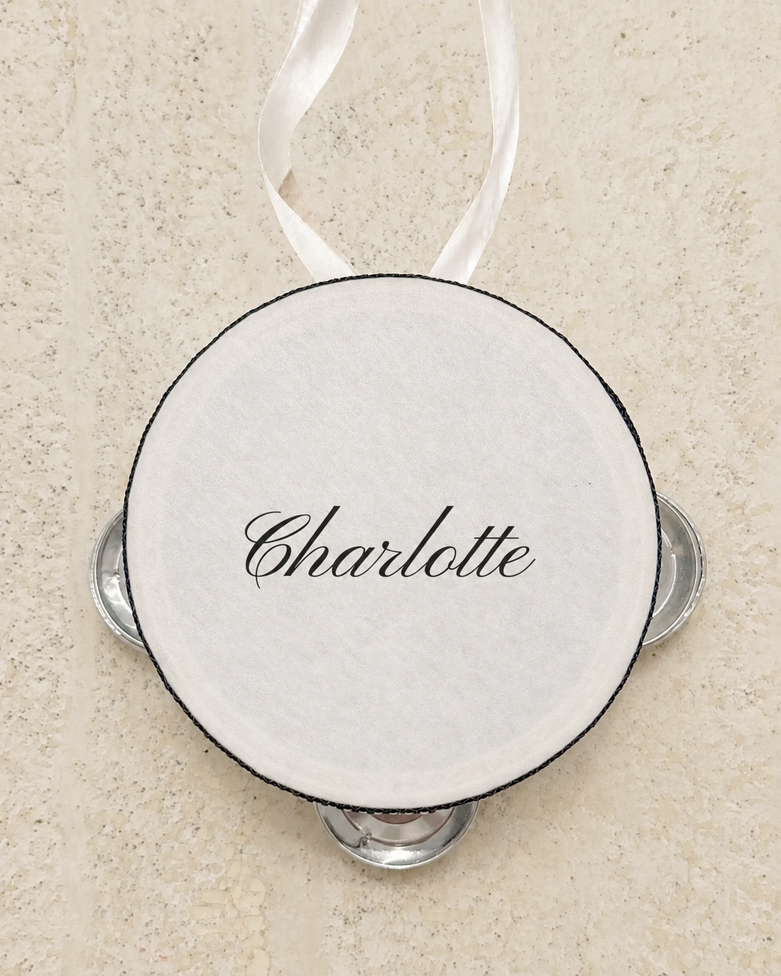 Personalised four inch tambourine place card tied with an ivory ribbon, on pale stone