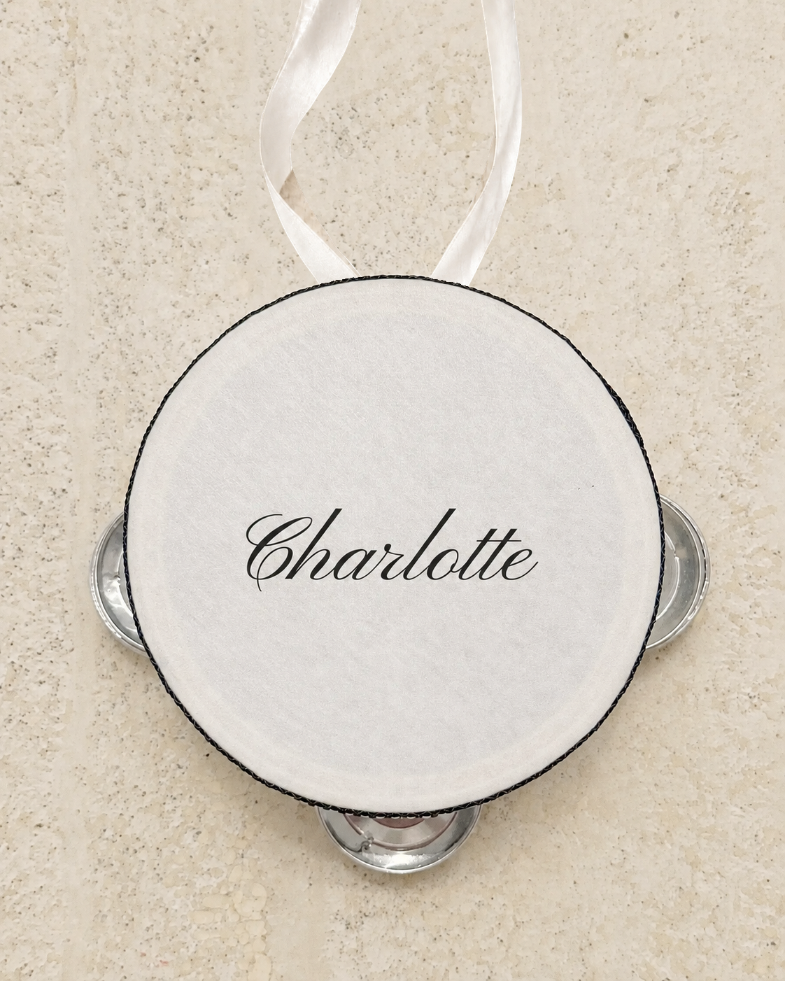Personalised four inch tambourine place card tied with an ivory ribbon, on pale stone