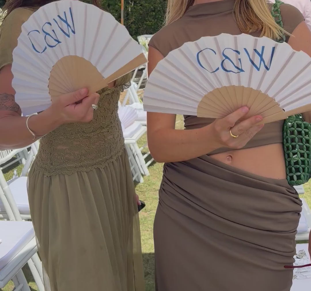 Two wedding guests holding personalised Solene fans printed with the couple's monogram at an outdoor ceremony